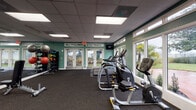 Located at 1658 S Hiawassee Rd in Orlando, Florida, this single-floor fitness center is a well-equipped exercise facility designed to support a full range of workout routines. The space features teal-painted walls and a drop-panel ceiling with recessed fluorescent lighting, creating a bright and energizing atmosphere throughout.

The cardio section is outfitted with multiple treadmills, elliptical machines, and a stationary bike, all positioned near large windows and glass double doors that flood the area with natural light and offer views of the surrounding outdoor greenery, including palm trees. A wall-mounted television is also present for entertainment during workouts.

The strength training area includes a multi-station cable and weight machine, a leg press, and a well-stocked dumbbell rack with a full range of free weights. Exercise balls, medicine balls, resistance bands, and additional accessories are neatly organized on dedicated storage racks, supporting functional and group fitness training.

Full-length mirrors line portions of the walls, enhancing the sense of space and allowing users to monitor their form. Rubber flooring throughout provides durability and safety underfoot. The facility also includes a water fountain and restroom access, ensuring user convenience. Overall, this fitness center offers a comprehensive and inviting environment for health and wellness activities.