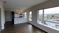 This single-floor space presents a clean, contemporary design centered around an open-concept living and kitchen layout. Dark hardwood flooring runs throughout, providing a warm contrast against the bright white walls and ceilings. Large windows along the living area wall flood the space with natural light and frame sweeping elevated views of the surrounding cityscape and hillside neighborhoods, making them a clear focal point of the unit.

The kitchen is thoughtfully integrated into the open floor plan via a peninsula-style counter with a white quartz or laminate surface, ideal for casual dining or food preparation. It is equipped with stainless steel appliances, including a refrigerator, microwave, and range, complemented by modern cabinetry featuring a combination of dark wood-toned and contemporary finishes. Pendant lighting hangs above the peninsula, while track lighting provides additional illumination over the cooking area.

The living space is generously proportioned, benefiting from the open layout and abundant window coverage. The overall ambiance is modern and airy, with a neutral color palette and elevated sightlines that enhance the sense of openness throughout.