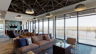 Located at 4190 W 17th Ave in Denver, Colorado, this upscale amenity lounge is a masterclass in contemporary design. The single-floor space is defined by a dramatic geometric lattice ceiling finished in dark tones, paired with stylish pendant lighting that creates a bold visual statement throughout the main lounge area. Floor-to-ceiling curtain wall windows run the length of the space, flooding the interior with natural light and offering sweeping panoramic views of the Denver skyline and surrounding landscape. A spacious outdoor terrace is accessible directly from the lounge, extending the living experience outdoors. The main seating area features generously sized sofas and leather accent chairs arranged around dark wood coffee tables, fostering an inviting and social atmosphere. A striking textured fireplace wall with a sleek linear fireplace serves as a focal point in one seating zone. The bar area is appointed with a central island, upholstered counter stools, mirrored globe pendant lights, warm wood cabinetry, and a mounted flat-screen display. A private conference or dining room offers a live-edge wood table, blue upholstered chairs, and a built-in media wall. A well-equipped catering kitchen with modern dark cabinetry and direct terrace access completes this thoughtfully designed communal space.
