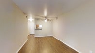 This single-floor unit offers a clean, functional layout designed for comfortable everyday living. The main living area is a welcoming, open space finished with rich hardwood-style flooring that adds warmth throughout. A ceiling fan with an integrated light fixture is centrally positioned, providing both air circulation and ambient lighting. Neutral-toned walls keep the space feeling bright and versatile, complemented by crisp white baseboards that frame the room.

A standout feature is the pass-through window that connects the kitchen to the living area, creating an open feel between the two spaces while maintaining a defined layout. The kitchen, visible through the pass-through, features warm wood-toned cabinetry and a countertop bar ledge, ideal for casual dining or entertaining. A hallway leads from the living area toward the entry door, providing a clear and practical flow through the unit.

The overall ambiance is clean and understated, with a neutral palette that serves as a versatile backdrop for a variety of interior styles. The hardwood-style floors lend a cohesive, polished look throughout, making this a straightforward and well-appointed single-floor space.