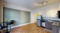 This single-floor unit offers a well-designed open-concept layout that seamlessly combines the living and kitchen areas into one cohesive space. The warm hardwood flooring runs throughout, lending a natural and inviting feel to the interior. A striking sage green accent wall adds a touch of color and character to the living area, complemented by crisp white trim and baseboards that frame the room cleanly.

The kitchen is efficiently appointed with stainless steel appliances, including a full-size refrigerator and dishwasher, paired with white upper cabinetry and dark countertops that create a modern contrast. A built-in track lighting fixture on the ceiling provides focused, adjustable illumination across the main living space.

The ceiling features classic decorative molding details that add architectural interest and a sense of traditional charm. A doorway visible in the background hints at additional space or storage beyond the main room. The overall ambiance is clean, bright, and functional, with a neutral palette punctuated by thoughtful design choices that make the space feel both practical and welcoming.