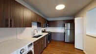 This single-floor unit offers a thoughtfully arranged layout that balances open living spaces with functional private areas. The living room is a standout feature, characterized by expansive floor-to-ceiling windows dressed with vertical blinds that frame urban skyline views and flood the space with natural light. Rich dark hardwood-style flooring runs throughout the main living and kitchen areas, creating a warm, cohesive aesthetic.

The kitchen is well-appointed with dark wood cabinetry extending to the ceiling for ample storage, white quartz countertops, a white tile backsplash, and a full suite of stainless steel appliances including a gas range, microwave, dishwasher, and refrigerator. An in-unit washer is also conveniently located within the kitchen area. A peninsula counter defines the transition between the kitchen and living space.

The bedroom features plush carpeting, a large window with city views, and access to closet space. The bathroom is finished in a clean, modern style with light wood-look tile flooring, a dark-stained vanity with a white countertop, a recessed medicine cabinet mirror, and a tiled tub-shower surround with a herringbone accent pattern.