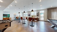 Located at 210 S Main St in Kannapolis, NC, this single-floor amenity space is designed as a modern, multifunctional clubroom that blends social gathering and recreational areas seamlessly. The interior is anchored by eye-catching chevron-patterned hardwood flooring that runs throughout, providing a warm and sophisticated foundation for the space.

The main lounge area features multiple high-top bistro tables with sleek modern bar stools, illuminated by a series of contemporary pendant lights and recessed ceiling fixtures. Large windows along the perimeter flood the room with natural light while offering views of the surrounding outdoor areas. A dedicated billiards zone occupies one end of the room, set against a bold amber accent wall adorned with vibrant pop-art prints that inject energy and personality into the space.

A well-appointed wet bar lines one wall, complete with dark wood cabinetry, a white quartz countertop, a undermount sink with a matte black faucet, and a draft tap — ideal for entertaining. Floating shelves above display decorative accents and greenery. A glass door adjacent to the wet bar provides direct access to an outdoor patio area with umbrella-shaded seating.

Additional seating vignettes include a live-edge communal table with teal metal stools, accented by wall-mounted artwork and sconce lighting. The overall ambiance is modern, vibrant, and inviting — well-suited for both casual socializing and community events.