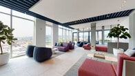 Located at 230 E Burnside St in Portland, Oregon, this single-floor amenity space offers a well-appointed, contemporary environment designed for both social gathering and productive work. The layout is expansive and open, anchored by floor-to-ceiling glass walls that frame sweeping panoramic views of the Portland skyline and surrounding cityscape, flooding the interior with abundant natural light throughout the day.

The main lounge area is thoughtfully arranged with modular seating in a vibrant palette of deep purple, coral red, and charcoal, positioned across multiple area rugs to define distinct conversation zones. Exposed structural concrete columns and bold navy ceiling beams contrast with the clean white surfaces, lending the space an industrial-modern character. Large-format tile flooring runs continuously throughout, reinforcing the sense of openness.

A fully equipped kitchen features a professional-grade stainless steel range, a generous island with neutral stone countertops, integrated cabinetry in muted gray, and a striking mosaic tile accent wall displaying a colorful, artistic pattern. A vivid pink faucet adds a playful design touch. A separate meeting area includes a solid wood table with matching chairs and direct city views through full-height glazing. Lush indoor plants are positioned throughout, softening the overall aesthetic.