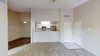 This single-floor residence offers an open-concept layout that connects the kitchen and living areas in a clean, functional design. The kitchen is equipped with stainless steel appliances, including a refrigerator and microwave, complemented by white cabinetry with wood-tone trim and a built-in overhead light. A white peninsula counter serves as a natural divider between the kitchen and the adjoining living space, providing additional prep and seating surface. The living area features neutral-toned carpet and benefits from vaulted ceilings that lend a sense of openness and height throughout. A pendant light fixture adds a subtle design accent above the dining or living zone. The entryway is finished with tile flooring, offering a durable transition from outside, and is flanked by a full-panel door alongside a closet for convenient storage. Warm beige walls and light-toned surfaces throughout create a cohesive, understated aesthetic. Ventilation is supported by ceiling vents positioned throughout the space. The overall layout is efficient and welcoming, well-suited for everyday living with a straightforward, low-maintenance interior design.