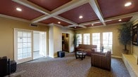 This single-floor space makes a strong first impression with its distinctive coffered ceiling, finished in rich burgundy panels framed by crisp white molding and accented with recessed lighting. The warm, neutral wall tones complement the patterned carpeting below, creating a cohesive and inviting atmosphere throughout. The main lounge area is furnished with comfortable upholstered sofas and armchairs arranged around a glass-top coffee table, offering a welcoming setting for gathering and conversation. Large multi-pane windows line one wall, allowing generous natural light to fill the room and enhance its open, airy feel. A tall potted plant adds a touch of greenery and life to the space. Adjacent to the main lounge, a kitchenette is visible through an open doorway, equipped with a full-size refrigerator and cabinetry, providing functional support for events or everyday use. French doors on the opposite side lead to additional rooms, suggesting versatility in how the space can be used. Overall, the space balances classic architectural detail with practical amenities, making it well-suited for both casual relaxation and organized gatherings.