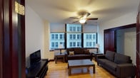 This 2-bedroom, 2-bathroom urban residence is situated on a single floor and offers a comfortable, functional layout ideal for city living. The living room serves as the heart of the home, featuring warm hardwood flooring that adds a welcoming, natural tone to the space. Large windows line the exterior wall, framing striking views of the surrounding city architecture and allowing natural light to fill the room throughout the day. A ceiling fan with integrated lighting adds both comfort and a practical touch to the living area. The living room is thoughtfully arranged with ample seating, a flat-screen television, and a central coffee table, creating an inviting space for relaxing or entertaining. The dark wood furnishings complement the light-toned walls and flooring, striking a balanced, cohesive aesthetic. The two bedrooms offer private retreats within the residence, while the two full bathrooms provide convenience for residents and guests alike. The overall ambiance of the space is urban and functional, with city views and natural light as defining characteristics that bring energy and a sense of connection to the surrounding environment.