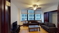 This 2-bedroom, 2-bathroom urban residence is situated on a single floor and offers a comfortable, functional layout ideal for city living. The living room serves as the heart of the home, featuring warm hardwood flooring that adds a welcoming, natural tone to the space. Large windows line the exterior wall, framing striking views of the surrounding city architecture and allowing natural light to fill the room throughout the day. A ceiling fan with integrated lighting adds both comfort and a practical touch to the living area. The living room is thoughtfully arranged with ample seating, a flat-screen television, and a central coffee table, creating an inviting space for relaxing or entertaining. The dark wood furnishings complement the light-toned walls and flooring, striking a balanced, cohesive aesthetic. The two bedrooms offer private retreats within the residence, while the two full bathrooms provide convenience for residents and guests alike. The overall ambiance of the space is urban and functional, with city views and natural light as defining characteristics that bring energy and a sense of connection to the surrounding environment.