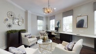 This single-floor space exudes a refined, contemporary elegance that balances functionality with sophisticated design. The main area serves as a seamlessly integrated office and lounge, anchored by a striking gold starburst chandelier that draws the eye upward to crisp crown molding and a smooth, light-toned ceiling. Rich hardwood flooring grounds the room with warmth and depth, complementing the neutral palette of soft grays and whites throughout.

Generous natural light pours in through multiple windows dressed with tailored gray Roman shades and white plantation shutters, creating a bright, inviting atmosphere. A built-in workstation with dark cabinetry and a clean white countertop runs along one wall, providing ample workspace framed by garden views. The seating arrangement features plush, cream-upholstered armchairs accented with textured throw pillows, gathered around a round glass-top coffee table with a gleaming gold geometric base. Curated artwork and sculptural accents lend an art-forward sensibility to the space. The overall ambiance is polished yet welcoming, thoughtfully designed to foster both productivity and relaxed conversation.