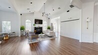 Dublin Terrace is a thoughtfully designed single-story community space that blends classic architectural details with a clean, modern aesthetic. The layout flows seamlessly from a welcoming entryway — featuring wainscoting, warm hardwood-style flooring, and elegant wall sconces — into a series of well-appointed gathering areas.

The main lounge is a standout feature, boasting soaring vaulted ceilings that amplify the sense of openness. Arched windows frame views of lush greenery, filling the space with abundant natural light. A black fireplace serves as the focal point, flanked by seating arrangements that include a neutral sofa, plush accent chairs, and a marble-top coffee table. A geometric pendant light adds a contemporary touch overhead.

Adjacent to the lounge, a dining area offers a marble oval table with upholstered chairs, positioned beneath wide windows with tree-lined views. A bookshelf adds a casual, inviting character to the room.

The kitchenette is finished with rich warm-toned cabinetry, granite countertops, tile flooring, and a window overlooking the outdoors. Additional flexible areas include an open multi-purpose room with a projection screen, suitable for presentations or events. Throughout, wainscoting, crown molding, and consistent flooring tie the entire space together cohesively.