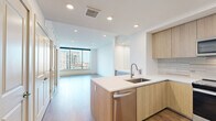 Located in the heart of Dallas's Uptown neighborhood, this well-appointed one-bedroom, one-bathroom residence offers a thoughtfully designed open layout with modern finishes throughout. Wide-plank hardwood flooring runs continuously across the living spaces, grounding the unit in warmth and cohesion.

The combined kitchen and living area anchors the unit, featuring a sleek, contemporary kitchen outfitted with flat-front cabinetry in a light wood finish, a tiled backsplash, stainless steel appliances including a range, microwave, dishwasher, and refrigerator, and a clean white quartz countertop. A peninsula provides additional prep and casual dining space.

Floor-to-ceiling windows with black frames and sliding glass door access to a balcony flood the living area with natural light while framing expansive views of the Dallas skyline and surrounding cityscape. The bedroom mirrors this feature with its own full-height window and glass door, a ceiling fan, and a bright, airy atmosphere.

The walk-in closet offers built-in shelving and rod storage for well-organized functionality. The bathroom presents a modern aesthetic with a floating wood vanity, gold-accented fixtures, a framed mirror, and tile flooring, with direct access to the walk-in closet. Clean white walls and recessed lighting complete the contemporary feel throughout.