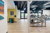 Located at 106 Yester Oaks Way in Greensboro, NC, this fitness center spans a generously sized single floor, thoughtfully designed to accommodate a full range of exercise and wellness activities. The facility is divided into several distinct functional zones, each with its own character while maintaining a cohesive modern aesthetic throughout.

The main exercise floor features light wood-look flooring and an open layout lined with True-brand cardio equipment, including treadmills, ellipticals, and stationary bikes. Large mirrors, wall-mounted televisions, and an exposed ceiling painted in deep navy blue contribute to an energetic yet polished atmosphere. Oversized ceiling fans and linear LED lighting ensure the space remains well-lit and comfortable. Glass double doors along one wall connect to an outdoor area, allowing natural light and outdoor views to enhance the environment.

A dedicated strength training zone features rubber flooring, power racks, cable machines, free weights, exercise balls, and large floor-to-ceiling mirrors. An adjacent multipurpose studio room offers a clean, open hardwood floor, a ceiling fan, interior windows, and wall-mounted artwork, making it suitable for group fitness classes or personal training sessions. The facility's yellow accent walls and curated artwork add warmth and visual interest throughout the space.
