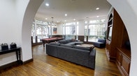 This single-floor amenity lounge is thoughtfully designed to offer both comfort and entertainment in an open, welcoming environment. The space is anchored by rich hardwood flooring that adds warmth and character throughout. An elegant arched entryway creates a natural transition into the main gathering area, where a large sectional sofa in neutral gray provides ample seating for socializing and relaxation.

A classic billiards table takes center stage in a dedicated game area, complemented by a modern pendant light fixture that adds a touch of refinement overhead. The space is framed by an impressive array of large, grid-paned windows on multiple walls, flooding the interior with natural light and offering pleasant views of mature trees and lush greenery outside. The bright, airy atmosphere is further enhanced by recessed ceiling lighting throughout.

Subtle design details — including wainscoting-style wall paneling, tasteful artwork, and carefully placed accent seating — contribute to an overall ambiance that is both polished and inviting. The open floor plan allows the space to feel expansive and versatile, suitable for both casual gatherings and more structured recreational use. Overall, this lounge strikes a refined balance between a functional community space and an elegant, comfortable retreat.