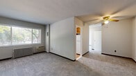 This 2-bedroom, 1-bathroom unit offers a straightforward and functional single-floor layout. The living area features neutral grey carpet, a ceiling fan with light fixture, and a wall-mounted air conditioning unit, with natural light filtering in through a window overlooking mature trees outside. The space flows toward a compact, galley-style kitchen outfitted with warm wood cabinetry, a gas range, a white refrigerator, a single-basin sink, and durable hardwood-style flooring. A window above the kitchen sink brings in additional natural light. The primary bedroom is generously sized, offering ample room for furnishings, grey carpet underfoot, and a window with outdoor views. The second bedroom is similarly appointed with carpet, neutral walls, and a window with natural light. Both bedrooms benefit from radiator heating. Two closets provide dedicated storage throughout the unit. The bathroom features a classic checkerboard black-and-white tile floor, a full soaking tub with tiled surround, a wood-vanity with an integrated sink, a frosted privacy window, and a vintage cast-iron radiator. The unit includes two closets for added storage convenience. Overall, the space presents a clean, no-frills aesthetic with practical finishes throughout.