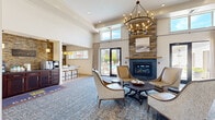 Located at 100 Holling Pl in Frederick, MD, this single-floor community amenity space offers a generously sized, thoughtfully designed interior that blends contemporary style with warm, inviting character. The centerpiece of the space is a sophisticated living and lounge area anchored by a floor-to-ceiling stacked stone fireplace with a wood mantel, complemented by soaring ceilings and an eye-catching wrought-iron chandelier. A chevron-patterned reclaimed wood accent wall adds texture and warmth, while plush seating arrangements and a patterned area rug create an inviting atmosphere for gathering and relaxation. Large glass doors open to outdoor views, flooding the lounge with natural light. Adjacent to the main lounge, a dedicated coffee bar station features dark cabinetry set against a stacked stone backsplash, offering a functional and stylish amenity. The kitchen area includes a substantial stone-topped island, stainless steel appliances, and matching dark wood cabinetry beneath a decorative tile accent wall branded with community signage. The hallways connect seamlessly to a well-organized package room with numbered shelving units for resident convenience. Throughout the space, hardwood-style flooring, recessed lighting, and pendant fixtures contribute to a polished, cohesive aesthetic.