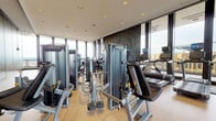 Located at 777-799 Main St in Hartford, Connecticut, this fitness center is a comprehensively appointed single-floor facility designed to support a full range of workout routines. The space is defined by its striking floor-to-ceiling glass curtain walls, which flood the interior with natural light and offer expansive views of the Hartford skyline and surrounding cityscape, creating an energizing atmosphere for exercise.

The main workout area features an extensive lineup of Matrix-brand strength and resistance machines arranged across light hardwood flooring, providing ample space to move between equipment. A row of Matrix elliptical trainers and treadmills is positioned directly in front of the panoramic windows, maximizing the outdoor views during cardio sessions. A decorative textured wall panel with an embossed laurel wreath motif serves as a bold architectural accent behind the cardio equipment.

A dedicated free weight and functional training zone features rubber flooring, adjustable benches, a dumbbell rack, medicine balls, and storage lockers for personal belongings. Full-length mirrors expand the sense of space and assist with form during workouts. Recessed ceiling lighting throughout ensures consistent illumination, complementing the abundant natural light. The entry area transitions seamlessly from an elegantly finished corridor with warm wood paneling and polished stone flooring, reflecting the building's upscale character.