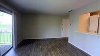 This single-floor space offers a straightforward and functional layout designed for comfortable everyday living. The main living area is open and uncluttered, presenting a blank canvas ready for personalization. Underfoot, rich dark hardwood-style flooring runs throughout, adding warmth and a modern aesthetic to the space. The walls are finished in a soft, neutral gray-beige tone that complements the flooring and contributes to a calm, cohesive atmosphere.

A white paneled entry door provides a clean focal point along one wall, flanked by a wall-mounted control panel — likely for climate or security — indicating that the space is equipped with practical modern conveniences. Ceiling vents are visible overhead, confirming a built-in HVAC system for year-round comfort. White baseboards trim the perimeter of the room, adding a crisp finishing detail that contrasts nicely with the darker flooring.

The space benefits from an even ceiling height that keeps the area feeling open and airy. The overall ambiance is minimal and move-in ready, with neutral finishes that offer versatility to suit a range of interior styles. Whether utilized as a living area or multi-purpose room, this space provides a solid, well-finished foundation.