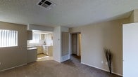 This single-story space is designed with an open, functional layout that connects the main living area to the kitchen and additional rooms. The living area is finished with wall-to-wall carpet in a neutral tone, contributing to a comfortable and understated ambiance. A textured popcorn ceiling runs throughout, and standard overhead HVAC vents ensure climate control.

The kitchen is accessible directly from the main living space through an open passthrough layout, featuring light-toned cabinetry, a double sink positioned beneath a window that allows natural light to filter in, and tile flooring that contrasts the carpeted living area. The kitchen is equipped with essential appliances and offers functional counter space along the perimeter.

A central structural column divides the living space from an interior doorway that leads to additional rooms, one of which features a built-in closet with visible shelving, offering practical storage solutions. The neutral beige walls throughout maintain a clean, cohesive look. The overall design is straightforward and utilitarian, suited for everyday living with an emphasis on functionality and ease of movement between spaces.
