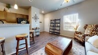 This single-floor home offers a thoughtfully designed layout that balances comfort and style. The open-concept living area is immediately welcoming, featuring rich hardwood-style flooring, vaulted ceilings with a ceiling fan, and generous natural light streaming through plantation-shuttered windows. A bold patterned accent chair, a sculptural solid-wood coffee table, and a neutral sofa anchored by a layered area rug create an inviting and well-curated gathering space. Leaning ladder shelves add decorative storage, while wall art and metallic accents contribute to the warm, eclectic aesthetic.

The kitchen is efficiently designed with dark espresso cabinetry, granite-style countertops, and full black appliance suite including a range, dishwasher, and microwave. A breakfast bar with bar seating connects the kitchen to the living space, making it ideal for both casual dining and entertaining.

The bedroom is a serene retreat, centered around a stately dark-wood bed frame and complemented by soft carpeting, matching nightstands, and ample natural light. A walk-in closet and additional closet storage provide excellent organization.

The full bathroom echoes the kitchen's design language with matching dark cabinetry, granite countertops, a single vanity, and a tub-shower combination. Consistent wood-look flooring and neutral tones unify the entire space.