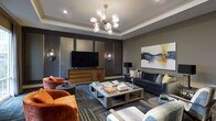 This single-floor space exudes a polished, contemporary elegance defined by thoughtful design and premium finishes. The living area serves as the centerpiece, showcasing a deep, moody color palette anchored by charcoal-toned walls and complemented by a tray ceiling with crisp white crown molding that adds architectural depth and a sense of refined structure.

A striking sputnik-style chandelier with amber glass globes and a brushed gold finish commands attention from above, casting a warm, ambient glow throughout the room. The space is anchored by a substantial wood-and-metal coffee table at the heart of the seating arrangement, surrounded by a plush slate-blue sofa, richly upholstered burnt-orange accent chairs, and layered textural cushions that invite comfort.

The entertainment wall is a standout feature, with custom paneled cabinetry in a muted grey finish flanking a large flat-screen television, accented by wall-mounted sconce lighting for a curated, boutique feel. A bold abstract artwork in navy, gold, and white adds a dynamic focal point to the adjacent wall.

Underfoot, a multi-tonal geometric area rug ties the varied furnishings together, while decorative accents including sculptural objects and curated books on the coffee table complete the layered, gallery-like ambiance of this sophisticated space.