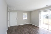 This single-story space presents a clean, straightforward layout designed for comfortable everyday living. The main living area is finished with rich, dark hardwood-style flooring that contrasts attractively against the neutral gray walls and bright white baseboards, creating a modern yet understated aesthetic. A textured popcorn ceiling adds a classic touch overhead, while a centrally placed air vent ensures efficient climate control throughout the space.

Natural light is a defining feature of this room, provided by a sliding glass door fitted with vertical blinds, which opens to an outdoor area and allows generous sunlight to stream across the flooring. A secondary window on the adjacent wall further brightens the interior, offering views to the outside. A white entry door with a deadbolt is conveniently positioned nearby, providing a clear and accessible entryway.

The overall ambiance is fresh and neutral, making the space highly adaptable for a variety of furniture arrangements and personal styles. The combination of durable flooring, ample natural light, and clean wall finishes gives this single-floor space a move-in-ready feel.