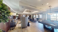 Located at 401 Northeast Mizner Boulevard in Boca Raton, FL, this sophisticated amenity lounge offers a well-appointed, open-plan layout spread across a single floor. The space is defined by wide-plank light wood flooring, tray ceilings with recessed lighting, and a neutral color palette of whites and warm grays that lend a clean, contemporary feel throughout.

The main gathering area features a comfortable lounge zone with upholstered seating, floor lamps, and greenery, creating an inviting atmosphere for relaxation. Adjacent to the lounge is a games area with two full-size billiards tables, illuminated by a statement crystal pendant fixture and benefiting from abundant natural light through multiple windows.

A central wet bar serves as the social hub of the space, showcasing rich dark wood cabinetry, a mosaic tile backsplash, stone countertops, a wall-mounted flat-screen TV, and pendant lighting with crystal detailing. Bar-height seating lines the island, making it ideal for entertaining.

A semi-private dining alcove with a dark wood table, upholstered chairs, and a built-in TV display provides an intimate setting for smaller gatherings or meetings. Dark wood doors and millwork accents appear consistently throughout, tying the design together with a refined, modern aesthetic.