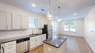 Located at 1200 River Dr in Belmont, NC, this well-appointed single-floor residence offers a thoughtfully designed layout with one bedroom, one bathroom, and a dedicated closet. The kitchen serves as a standout feature, showcasing white shaker-style cabinetry, dark granite countertops, a classic white subway tile backsplash, and a full suite of stainless steel appliances including a range, microwave, dishwasher, and refrigerator. A central island with a matching granite surface provides additional prep space and casual seating potential. Pendant lighting and recessed fixtures illuminate the space, while light hardwood-style flooring ties the kitchen and main living areas together seamlessly. The bedroom is a comfortable retreat, finished with soft wall-to-wall carpeting, a ceiling fan, and a window that allows natural light and outdoor views. It connects to a generously sized walk-in closet outfitted with wire shelving systems for organized storage. The bathroom is clean and modern, featuring hardwood-style flooring, a gray vanity with a dark granite countertop, an integrated sink, a framed mirror, and contemporary lighting above. The overall aesthetic throughout the space is bright, fresh, and move-in ready, with a neutral color palette that creates a cohesive and inviting atmosphere.