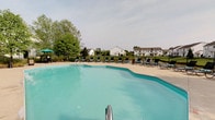 Located at 6340 Saddler Way in Canal Winchester, Ohio, this single-level community amenity area centers around a standout outdoor swimming pool designed for relaxation and recreation. The pool features a gently curved, freeform shape filled with clear, inviting water, making it a focal point of the outdoor space. A broad concrete pool deck surrounds the water, offering ample room for movement and socializing. A generous arrangement of lounge chairs lines the perimeter of the deck, providing dedicated spaces for sunbathing and leisure. A shaded seating area with a patio umbrella is also present, offering a cooler retreat on warm days. The pool area is enclosed by a black iron fence, providing both security and an open, airy feel that preserves views of the surrounding landscape. Lush mature trees and green open lawn areas frame the space, contributing to a serene, park-like atmosphere. In the background, neighboring residential buildings are visible, underscoring the community-oriented nature of this amenity. The overall setting is open, well-maintained, and thoughtfully laid out to encourage outdoor enjoyment in a welcoming neighborhood environment.