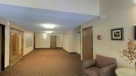 This single-floor corridor presents a straightforward, functional layout designed for efficient movement and accessibility. The hallway features warm-toned, patterned carpet flooring that runs the length of the space, complemented by light-colored walls that contribute to a clean, neutral aesthetic. Flush-mounted rectangular ceiling light fixtures provide consistent overhead illumination throughout the corridor, while a textured ceiling adds subtle architectural detail.

Multiple wood-toned doors line the hallway, each fitted with standard hardware, providing access to various rooms or units along the passage. A small window near one of the doors allows a modest amount of natural light to supplement the artificial lighting. Near the entrance area, what appears to be a mailbox or notice panel is mounted on the wall, suggesting a communal or residential building setting.

A padded, upholstered armchair is positioned near the end of the corridor, offering a small seating area for occupants or visitors. A wall-mounted fire safety device is also visible, indicating the space adheres to standard safety requirements. Overall, the corridor maintains a practical, well-maintained environment suited for daily use.