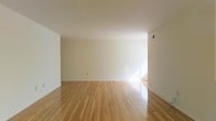 This single-floor space presents a clean and minimalist interior defined by smooth, warm-toned hardwood floors that run throughout the open living area. The floors are well-maintained and reflect natural light, contributing to the bright and airy atmosphere of the room. The walls are finished in crisp white, creating a neutral backdrop that enhances the sense of openness and allows for flexible use of the space.

The ceiling is smooth and painted white, with a standard HVAC vent integrated cleanly into the layout, ensuring functional climate control without disrupting the minimal aesthetic. Natural light enters from what appears to be a window or opening toward the far end of the room, casting a soft, warm glow across the floor and walls.

The overall layout is uncluttered and versatile, making it well-suited for a variety of configurations. The combination of light hardwood flooring and white walls gives the space a timeless, neutral character. The absence of decorative elements keeps the focus on the architectural simplicity and quality of the foundational finishes, offering a solid canvas for any intended use.