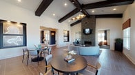 This thoughtfully designed single-floor clubhouse offers a range of distinct spaces unified by a contemporary aesthetic. The main gathering area commands attention with soaring vaulted ceilings accented by bold dark wood beams and recessed lighting. A floor-to-ceiling stacked stone fireplace serves as a dramatic focal point, complemented by a wall-mounted television and a curved sectional sofa arrangement on a patterned area rug — creating an inviting lounge setting. Adjacent to this area, round bistro tables and modern chairs provide casual seating throughout the open common space, which flows over warm large-format tile flooring.

The kitchen bar area features sleek dark cabinetry, striking veined stone countertops, and a full subway-tiled accent wall, outfitted with appliances and bar seating — ideal for a coffee station or social gathering spot. Curated recessed art niches with built-in lighting add gallery-quality detail to the walls throughout.

A dedicated business center offers workstations set against a bold geometric-patterned feature wall beneath a vibrant orange coffered ceiling. A separate lounge room continues the orange accent palette, with a coffered ceiling, plush seating, carpet flooring, and generous natural light streaming through multiple windows — completing this versatile and well-appointed communal facility.