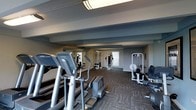 This fitness center is a fully appointed exercise facility designed to meet a variety of workout needs. The space is laid out on a single floor with a practical, open floor plan that allows for easy movement between equipment stations. Underfoot, durable patterned carpet tiles provide both comfort and traction throughout the room.

The cardio section is prominently featured and includes multiple elliptical trainers and treadmills, each equipped with individual digital display consoles for tracking workout metrics. Toward the rear and sides of the room, a selection of strength-training cable machines and multi-use weight stations offer options for resistance and muscle-building exercises. Additional seated workout equipment is also positioned throughout the space.

Overhead, recessed structural beams define the ceiling and contribute to the room's architectural character, while flush-mounted fluorescent lighting ensures the entire area is well-lit. A large mirror along one wall enhances the sense of space and allows users to monitor their form during exercise. Natural light filters in through a window at the far end of the room, adding a welcoming brightness. The overall ambiance is clean, functional, and focused, making this fitness center a practical and well-organized resource for daily exercise.