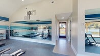 This single-level fitness facility is thoughtfully designed to support an active lifestyle, combining functional amenities with a clean, modern aesthetic. The centerpiece of the space is a generously sized gym area featuring vaulted ceilings with recessed lighting and a bold ring-shaped pendant fixture. Teal and white horizontal stripes run along the walls, complemented by bold motivational lettering, including a prominent "DON'T QUIT" wall graphic, creating an energizing atmosphere throughout. The gym is equipped with a comprehensive range of fitness equipment, including treadmills, ellipticals, a rowing machine, a multi-station cable machine, and a full dumbbell rack. A large floor-to-ceiling mirror spans one wall, enhancing the sense of space and allowing users to monitor their form. Rubber flooring throughout the workout area provides durability and comfort underfoot. Windows along the gym walls invite natural light and offer views to the exterior. An accessible restroom is also included, featuring light-toned tile flooring, a floating white vanity with drawers, teal subway tile backsplash, and ADA-compliant grab bars. A dedicated laundry room rounds out the facility, housing multiple commercial-grade washer and dryer units set against light tile flooring and neutral walls.