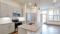 This single-floor residence showcases a thoughtfully designed open-concept layout with a seamless flow between the kitchen and living areas. The kitchen serves as the centerpiece, featuring a large island with granite countertops, an undermount sink with a pull-down faucet, and stainless steel appliances including a refrigerator, range, microwave, and dishwasher. Gray shaker-style cabinetry extends to the ceiling, maximizing storage, while a white subway tile backsplash adds a clean, contemporary touch. Geometric gold-toned pendant lights hang above the island, contributing a modern aesthetic. Wood-look flooring runs consistently throughout the unit, lending warmth and continuity. The bedrooms are bright and airy, each equipped with a ceiling fan and windows that bring in natural light. The primary bathroom is finished with large-format marble-look tile on both the floors and tub surround, complemented by a granite-topped vanity and a spacious walk-in closet featuring multiple shelving tiers and hanging rods. The overall design palette of neutral whites and grays creates a clean, contemporary ambiance throughout the space.