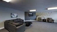 This single-floor space offers a comfortable and thoughtfully designed lounge area that blends modern aesthetics with welcoming touches. The main living area is anchored by a bold dark accent wall, against which a flat-screen TV is mounted, creating a natural focal point for the room. Soft, neutral-toned carpet runs throughout, adding warmth underfoot and complementing the cool grey tones of the walls and textured ceiling.

Seating is arranged to encourage relaxed gatherings, featuring a plush dark sofa, an additional upholstered chair with a bold pattern, and a sleek geometric black coffee table that adds a contemporary edge to the space. A decorative armchair with a side table and lamp occupies a cozy corner, providing a quieter spot for reading or unwinding.

One of the most distinctive features of the space is the hand-painted tree mural that extends across the lighter accent wall, with stylized green leaves adding an organic, artistic element that softens the room's otherwise modern palette. A framed abstract artwork on the opposite wall further reinforces the creative character of the interior. Overhead fluorescent lighting ensures the space remains bright and functional. Overall, the room exudes a relaxed yet polished ambiance, suitable for lounging, entertainment, or social gatherings.