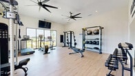 The Yardly Stoneridge fitness center at 322 W Melissa Rd in Melissa, TX offers a thoughtfully designed, single-floor workout facility. The space features light wood-style flooring and crisp white walls that create a clean, energizing atmosphere. Large floor-to-ceiling windows line the perimeter, flooding the gym with abundant natural light and providing open views of the surrounding outdoor landscape. Multiple oversized ceiling fans ensure effective air circulation throughout the space.

The facility is comprehensively equipped to support a wide range of fitness routines. Cardio options include treadmills, an elliptical, and a stationary cycle, each featuring individual digital screens. Strength training is well represented with a multi-function cable machine, a power rack with a bench press, a rowing machine, and an adjustable weight bench. A dedicated free weights area provides a full dumbbell rack, medicine balls, stability balls, and TRX suspension straps, all neatly organized on storage racks. Wall-mounted flat-screen televisions are positioned throughout for entertainment during workouts. A large wall mirror enhances the sense of space and supports form monitoring. The Yardly Stoneridge branding is prominently displayed, reinforcing the community identity of this well-appointed amenity.
