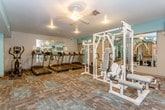 This fitness center offers a comprehensive workout environment spread across a single, open floor. The space is defined by its patterned carpet flooring with rubber mat overlays in key workout zones, neutral gray walls accented by teal highlights, and ceiling fans paired with recessed and pendant lighting throughout. The main workout area is generously sized, accommodating a wide variety of equipment without feeling cramped. A dedicated cardio section houses a row of treadmills and an elliptical machine, complemented by large mirrors along the wall to enhance the sense of space. The strength training area features a multi-station cable machine, several weight benches, and rubber-matted flooring ideal for free weight exercises. A full dumbbell rack lines one wall, offering an extensive range of weights for varied training needs. Barbell plates and additional benches are positioned throughout the open floor for functional and compound lifts. The entry area includes a wall of individual lockers, a posted fitness center rules board, a fire extinguisher, and a bulletin board, providing a practical and organized welcome to the space. Natural light filters in through a window and exterior door, while wall-mounted televisions add an entertainment element to the workout experience.