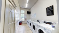 Located at 1001 Bear Island Rd in Summerville, South Carolina, this shared laundry facility is a well-appointed, single-floor amenity space designed to serve the needs of the community. The room is bright and clean, with neutral-toned walls, crown molding detailing, and light tile flooring that create a polished, welcoming atmosphere.

Along one side of the space, a row of commercial-grade front-load washing machines provides ample capacity for residents. Opposite the washers, stacked dryer units maximize the use of vertical space, offering an efficient and organized layout. The room is well-lit by overhead fluorescent lighting, complemented by natural light streaming through an arched window that overlooks a lush, green outdoor landscape.

A wall-mounted flat-screen television adds a touch of comfort, allowing residents to pass the time conveniently while doing laundry. Instructional signage is posted on the walls to guide users through proper machine operation. A fire extinguisher is also readily accessible, reflecting attention to safety. Two entry doors — one on each end of the room — ensure easy access and smooth flow of foot traffic. The overall ambiance is clean, functional, and thoughtfully designed to make the laundry experience as convenient as possible.
