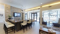 This contemporary single-floor space is designed to impress, blending modern aesthetics with a warm, inviting atmosphere. The centerpiece of the lounge is a sleek, dark bar-height counter surrounded by cream upholstered high-back stools, creating an ideal setting for casual gatherings or collaborative work. A textured stone accent wall adds a striking natural element to the space, complemented by a wall-mounted flat-screen television that enhances the room's entertainment appeal. Overhead, a cluster of polished spherical pendant lights serves as a dramatic focal point, casting a warm ambient glow that pairs beautifully with the recessed cove lighting integrated into the tray ceiling. Light-toned wood-style flooring runs throughout, lending a clean and cohesive look to the interior. Floor-to-ceiling glass doors along the far wall flood the space with generous natural light and offer clear views to the outdoors, creating a seamless connection between interior and exterior. An adjacent seating area with patterned upholstered furniture provides a relaxed lounge option, completing this well-appointed and thoughtfully designed communal environment.