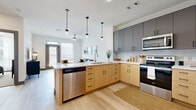 This thoughtfully designed single-floor residence blends modern aesthetics with functional living. The open-concept kitchen is a standout feature, showcasing a two-tone cabinet design pairing warm natural wood lower cabinets with slate-grey uppers, complemented by white quartz countertops, a subway tile backsplash, and a full suite of stainless steel appliances including a dishwasher, range, and microwave. A peninsula with bar seating and sleek pendant lighting makes it ideal for both everyday meals and entertaining. The kitchen flows seamlessly into the living area, where light hardwood-style flooring, large windows with abundant natural light, and a glass-paneled entry door create an airy, welcoming atmosphere. The living room offers a comfortable layout with generous wall space and direct access to the outdoors. The bedroom is carpeted for warmth and comfort, featuring a ceiling fan, large windows with sheer curtains, and ample room for full-sized furnishings. An expansive walk-in closet with multiple hanging rods and built-in shelving provides excellent storage. The en-suite bathroom impresses with a long double-sink vanity in natural wood tones, a wide mirror, white quartz countertops, and a glass-enclosed walk-in shower with tile surround. Throughout the space, neutral tones and clean lines establish a cohesive, contemporary ambiance.