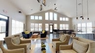 This single-story amenity space is defined by its dramatic vaulted ceilings and an open, airy layout that seamlessly connects multiple functional zones. The main lounge area features soaring cathedral ceilings with clerestory windows that flood the interior with natural light, complemented by a ceiling fan, industrial-style pendant lighting, and views of an outdoor pool. Comfortable seating arrangements include navy sofas accented with yellow throw pillows and striped armchairs grouped around coffee tables on woven rugs, creating inviting gathering spots.

The fully equipped kitchen is a standout feature, showcasing a striking stacked-stone accent wall that extends to the ceiling, open wooden shelving, granite countertops, stainless steel appliances, white cabinetry, and a peninsula with woven barstool seating. A vintage-style popcorn machine adds a playful touch near the kitchen entrance.

A dedicated game and dining area offers high-top wooden tables with industrial barstools alongside a shuffleboard table, all set against large windows overlooking the pool area. A built-in workstation, recessed into a textured accent wall and equipped with computers and a printer, provides a practical business center. Throughout, large-format tile flooring ties the space together cohesively.