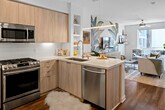 Located at 1901 E Dyer Rd in Santa Ana, CA, this modern single-floor unit offers a thoughtfully designed open-concept layout that seamlessly connects the kitchen, living, and sleeping areas. The kitchen is a standout feature, outfitted with light wood flat-front cabinetry, a white subway tile backsplash, stainless steel appliances including a gas range, microwave, dishwasher, and French door refrigerator, and a peninsula with quartz countertops that doubles as a breakfast bar with seating for two. Rich hardwood-style flooring flows throughout the entire space, adding warmth and continuity. The living area is full of personality, anchored by a bold tropical botanical mural that serves as a dramatic focal point behind the media console. A ceiling fan, natural light from a large window, and access to a balcony enhance the room's airy feel. The bedroom is well-appointed with a decorative chandelier, patterned wallpaper accent wall, and a window allowing natural light. The bathroom features a light wood vanity with a white quartz countertop, a large framed mirror, and dark tile flooring. Open shelving throughout adds functional display space, completing this stylish and well-equipped home.