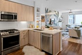 Located at 1901 E Dyer Rd in Santa Ana, CA, this modern single-floor unit offers a thoughtfully designed open-concept layout that seamlessly connects the kitchen, living, and sleeping areas. The kitchen is a standout feature, outfitted with light wood flat-front cabinetry, a white subway tile backsplash, stainless steel appliances including a gas range, microwave, dishwasher, and French door refrigerator, and a peninsula with quartz countertops that doubles as a breakfast bar with seating for two. Rich hardwood-style flooring flows throughout the entire space, adding warmth and continuity. The living area is full of personality, anchored by a bold tropical botanical mural that serves as a dramatic focal point behind the media console. A ceiling fan, natural light from a large window, and access to a balcony enhance the room's airy feel. The bedroom is well-appointed with a decorative chandelier, patterned wallpaper accent wall, and a window allowing natural light. The bathroom features a light wood vanity with a white quartz countertop, a large framed mirror, and dark tile flooring. Open shelving throughout adds functional display space, completing this stylish and well-equipped home.