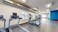Located at 4400 Redhawk Dr SE in Rochester, Minnesota, this well-equipped exercise room offers a generous, single-floor layout designed to accommodate a wide range of fitness activities. The space is thoughtfully organized into distinct workout zones, maximizing usability for both cardio and strength training.

Along one wall, a row of treadmills and elliptical trainers — including Precor equipment — are positioned beneath large windows that bring in natural light and offer views of the surrounding neighborhood. Exposed industrial-style ductwork runs along the ceiling, adding a utilitarian aesthetic that complements the fitness environment.

The strength training area features cable machines, weight racks, benches, and a full dumbbell rack, providing comprehensive options for resistance-based workouts. A vibrant blue accent wall anchors the functional training zone, which includes TRX suspension straps, medicine balls, plyo boxes, and a multi-purpose training rig — ideal for dynamic, high-intensity exercises.

Durable rubber and interlocking foam flooring covers the space, providing safety and support underfoot. Flush-mounted ceiling light fixtures ensure bright, even illumination throughout. Multiple windows along the perimeter walls fill the room with daylight, creating an energizing atmosphere. The overall design balances practicality with a motivating environment suited for a full-body fitness routine.