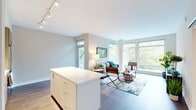 This thoughtfully designed single-floor residence offers a seamless blend of modern finishes and an abundance of natural light. The open-concept layout connects the kitchen and living areas, creating an airy, cohesive flow throughout the space. Rich hardwood flooring runs through the main living areas, anchoring the contemporary design.

The kitchen is outfitted with stainless steel appliances, sleek wood-toned cabinetry extending to the ceiling, a white quartz countertop, and a central island on casters — providing both additional prep space and flexibility. Track lighting illuminates the workspace efficiently.

The living room is a standout feature, wrapped in expansive floor-to-ceiling windows that frame lush, verdant tree canopy views and flood the room with natural light. A structural column adds an architectural accent without disrupting the open feel, while a sliding glass door offers access to an outdoor balcony area.

The primary bedroom continues the theme of generous natural light through a wide window bank overlooking the greenery, complemented by soft carpeting. A second bedroom provides comfortable accommodations with a built-in closet with shelving. The bathroom features a clean, neutral palette with a modern vanity, white quartz countertop, tile flooring, and a shower-tub combination.