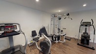 This single-floor fitness room offers a comprehensive and functional workout environment suited for a range of exercise routines. The space is finished with warm wood-look flooring throughout, providing a clean and durable surface that complements the room's bright, neutral-toned walls. Recessed ceiling lighting ensures even illumination across the entire area, while large windows dressed with vertical blinds allow natural light to filter in, contributing to an open and energizing atmosphere. A wall-mounted air conditioning unit provides climate control for year-round comfort during workouts.

The room is equipped with an impressive array of exercise machines to support both cardiovascular and strength training goals. Cardio equipment includes multiple treadmills, a stationary recumbent bike, elliptical trainers, and a stair-stepper, offering diverse options for aerobic exercise. For strength training, a multi-station cable and weight machine provides a wide variety of resistance exercises, accommodating users of different fitness levels. The equipment is thoughtfully arranged to allow sufficient space for movement and safe use of each machine. Posted guidelines on the wall help users navigate the space effectively. Overall, the fitness room presents a well-organized, practical, and accessible facility designed to support a complete workout regimen.