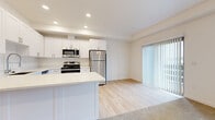 This well-appointed single-floor residence offers a clean, contemporary design throughout. The open-concept kitchen serves as the heart of the home, outfitted with crisp white shaker cabinetry, light quartz countertops, and a full suite of stainless steel appliances including a gas range, over-the-range microwave, dishwasher, and refrigerator. A generously sized peninsula with an undermount sink provides both additional prep space and a natural division between the kitchen and the adjoining living area. Light hardwood flooring runs throughout the kitchen and living space, lending warmth and continuity to the layout. The living area benefits from abundant natural light, with a large window and vertical blind-covered sliding glass door that opens to the outdoors. The bedrooms are carpeted for comfort and feature neutral walls, window blinds for light control, and views of the surrounding neighborhood. At least one bedroom includes a closet with built-in shelving and hanging storage. The overall aesthetic is bright and neutral, with recessed lighting, clean white walls, and consistent trim work throughout, creating a move-in-ready space that is both functional and inviting.