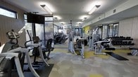 This well-appointed fitness center offers a comprehensive workout environment designed to accommodate a wide range of exercise needs. The space is outfitted with an impressive array of cardio equipment, including stationary bikes and elliptical trainers, alongside a full selection of strength training machines from leading brands. Floor-to-ceiling mirrors line the walls, enhancing the sense of openness and allowing users to monitor their form throughout their workout. Large windows along one side of the facility flood the space with natural light, creating an energizing atmosphere while offering views of the surrounding greenery. The interior design is cohesive and contemporary, featuring a distinctive hexagonal motif carried through both the decorative wall panels and the patterned flooring, which combines soft grey and yellow tones for a modern, vibrant aesthetic. Overhead, bright linear lighting ensures the entire facility is well-lit throughout the day. A ceiling fan adds to the comfort of the environment during intense workouts. The layout is thoughtfully organized, with cardio equipment positioned near the windows and strength machines arranged to allow ample circulation space, making the fitness center both functional and visually appealing.