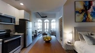 This single-floor residence showcases a thoughtfully designed open-concept layout that connects the kitchen, dining, and living areas in one continuous flow. The kitchen is equipped with stainless steel appliances, including a full-size refrigerator, and features clean white cabinetry that contributes to a bright, modern aesthetic. A warm-toned hardwood dining table anchors the central space, creating a natural transition point between the kitchen and living room.

The living area is a standout feature, distinguished by a striking set of tall arched windows that flood the space with abundant natural light and offer views of the surrounding urban environment. Exposed ductwork along the ceiling adds an industrial character that contrasts appealingly with the otherwise refined interior. The living room is comfortably furnished with a contemporary sofa and area rug, framed by the dramatic window backdrop.

A decorative patterned door panel hints at additional rooms beyond, while a side table with a lamp adds warmth near the entryway. The overall ambiance balances industrial and modern residential elements, creating an inviting and visually dynamic living environment.
