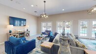 Located in Ponte Vedra Beach, Florida, this single-story clubhouse is designed with a vibrant coastal aesthetic that sets a welcoming tone throughout. The main gathering area is generously proportioned, featuring light wood-look flooring, recessed lighting, and statement geometric pendant fixtures that anchor the room with modern style. Comfortable seating arrangements — including plush blue velvet sofas, a tufted gray sectional, and accent chairs in rich teal and cobalt tones — are centered around natural live-edge wood coffee tables, reinforcing the organic, beach-inspired theme. A large wall-mounted flat-screen TV and a barn-wood media console complete the lounge area. Multiple sets of white-framed French doors flood the space with natural light and provide direct views of and access to the outdoor area, likely a pool. A decorative console table adorned with teal vases and a colorful framed mirror adds an elegant accent corner. The kitchen area features white cabinetry, granite countertops, stainless steel appliances including a refrigerator and microwave, and a breakfast bar-style counter that opens to the main gathering space. Coastal artwork throughout ties the design together, creating a cohesive, relaxed atmosphere ideal for community gatherings.